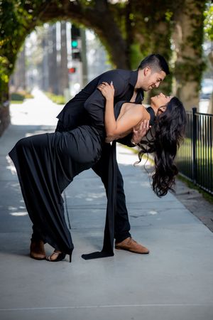 Couple in black formal wear performing a dramatic dip pose on an outdoor sidewalk with lush green trees.
