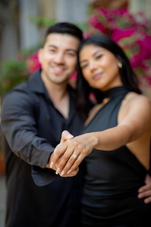 Newly engaged couple proudly displaying diamond engagement ring toward camera with pink flowers in background.