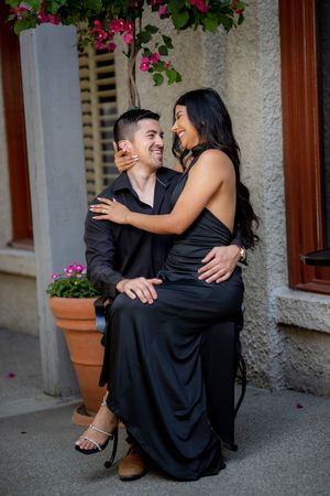 Happy couple in black outfits laughing together on steps adorned with colorful potted flowers and plants.