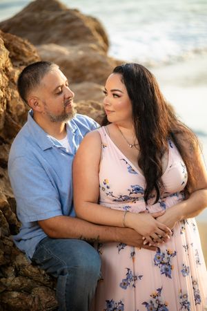 Couple gazes lovingly at each other while seated against rocky coastal backdrop with ocean waves behind them.