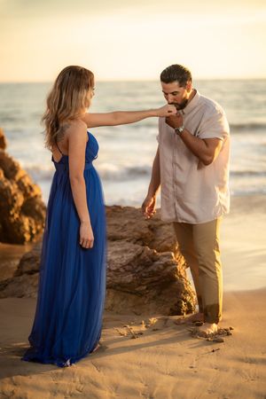 Man gallantly kisses womans hand on sandy beach at sunset, woman wearing elegant blue gown.