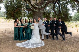 Wedding party poses playfully under a large oak tree with bridesmaids in emerald green dresses.