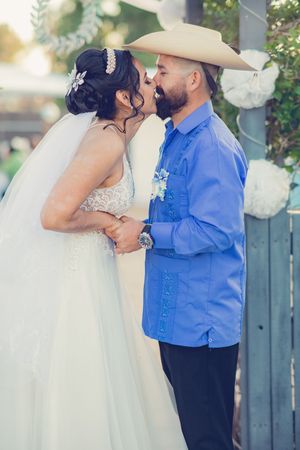Bride and groom in a blue shirt and cowboy hat share a kiss at their outdoor wedding ceremony.