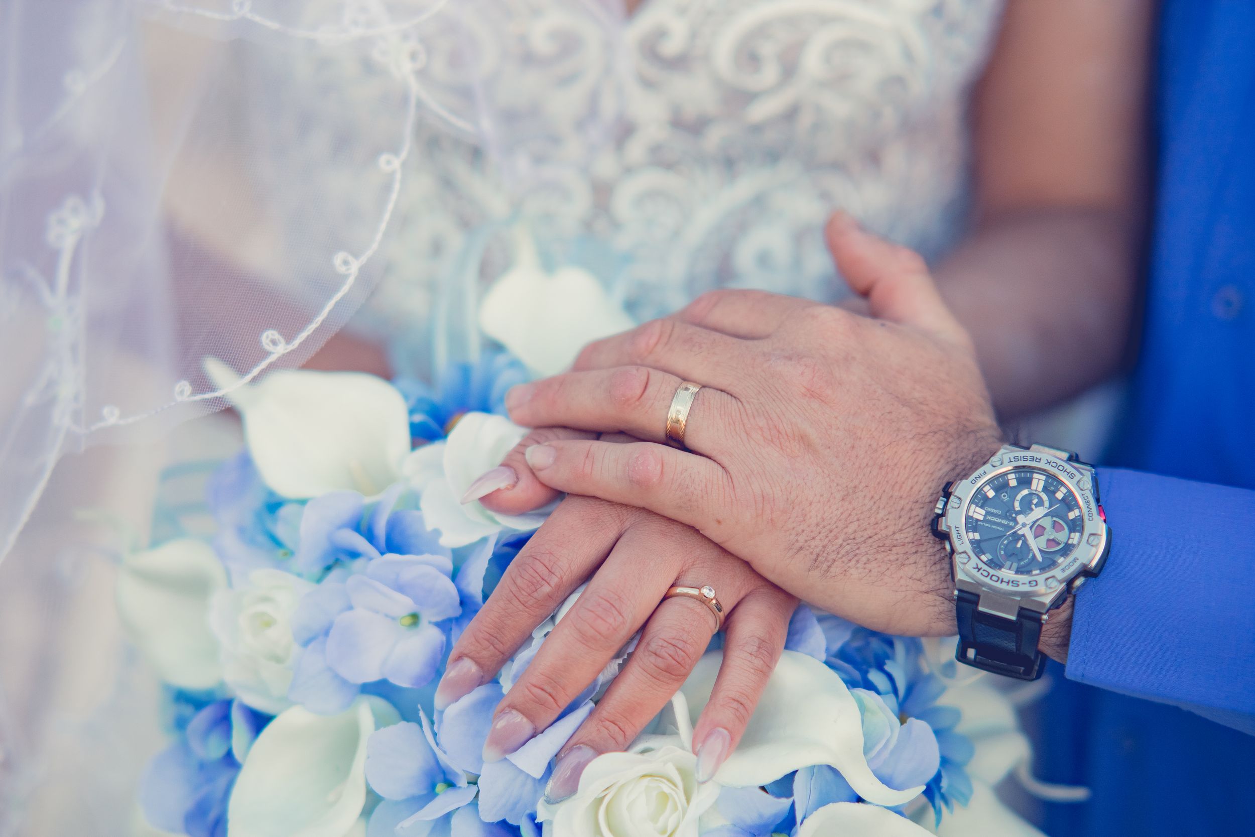 Newlyweds display their wedding rings over a bouquet of blue hydrangeas and white roses.