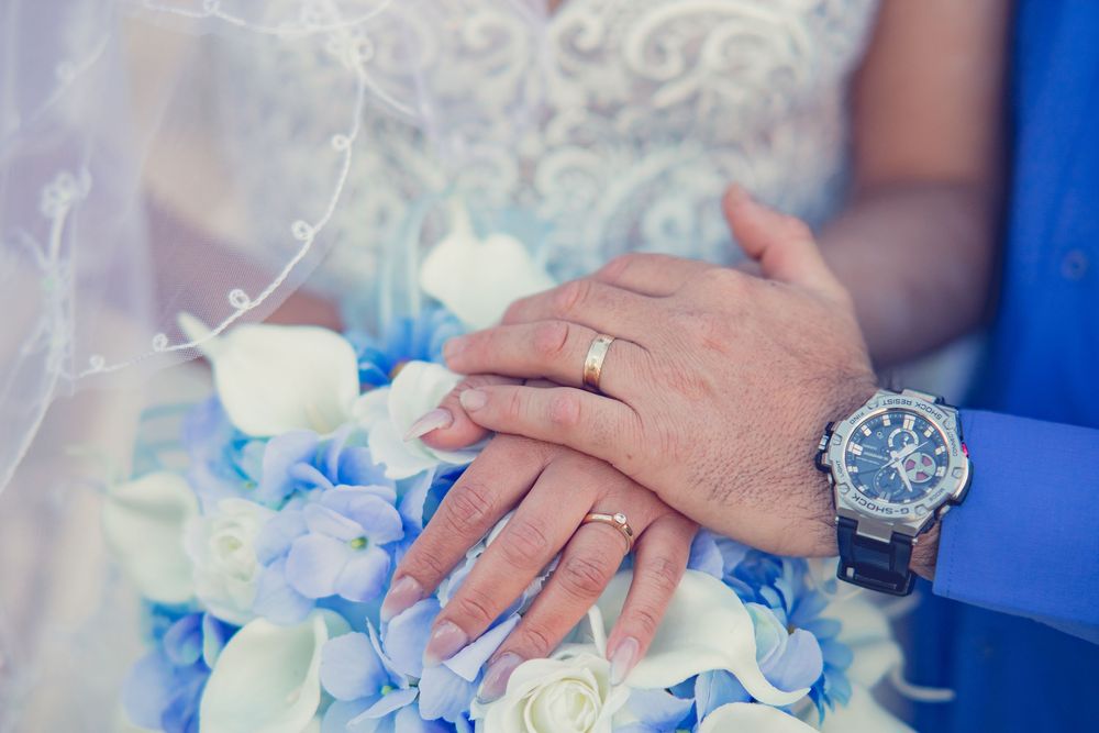 Newlyweds display their wedding rings over a bouquet of blue hydrangeas and white roses.