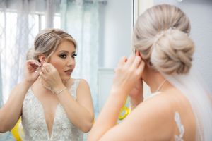 A bride in a lace wedding gown putting on earrings while looking in a mirror before her wedding.