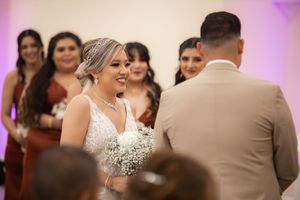 Smiling bride in lace gown holds white bouquet during indoor wedding ceremony with bridesmaids in burgundy dresses.