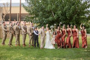 Large wedding party poses outdoors with groomsmen in tan suits and bridesmaids in rust-colored gowns under a tree.