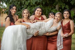 Four bridesmaids in rust satin dresses playfully carry smiling bride in white lace gown outdoors.