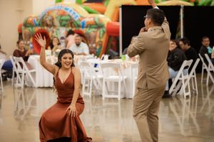 Woman in rust satin gown dances energetically with man in tan suit at wedding reception with colorful bounce house.