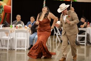 Couple dances joyfully at wedding reception, man in tan suit and cowboy hat, woman in flowing rust gown.