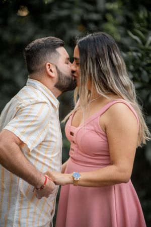Couple shares a romantic kiss outdoors, man in striped shirt, woman in pink dress.
