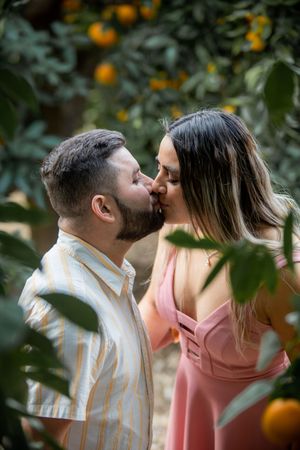 Couple shares a tender kiss among orange trees, man in striped shirt, woman in pink dress.