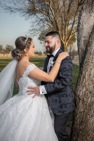 Bride and bearded groom in navy tuxedo share a romantic moment leaning against a tree at sunset.