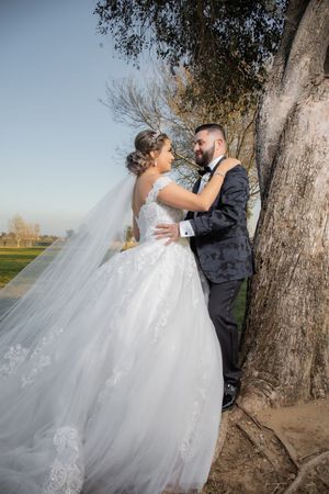 Bride and groom share a romantic embrace near a large tree outdoors during their wedding day.
