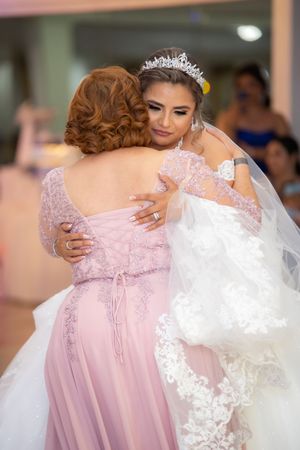 Bride in a white lace gown and tiara shares a heartfelt hug with a woman in a pink dress.