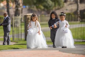 Three young flower girls in white dresses walking along a path at an outdoor wedding ceremony.