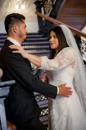 Bride in lace veil and groom in black suit sharing tender moment on ornate staircase.