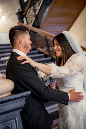 Smiling bride in lace wedding dress embracing groom on elegant staircase during first look.