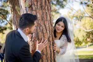 Bride in white lace gown and veil peeks around a tree at groom in black suit during outdoor wedding photos.