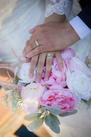 Newlyweds display wedding rings resting hands on pink and white peony bridal bouquet.
