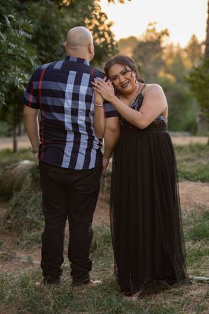 Woman in black formal gown smiles at camera while leaning on man in plaid shirt at sunset.