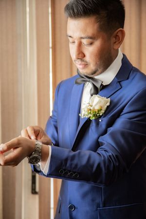 A groom in a blue suit with a white boutonniere checking his watch before the wedding ceremony.