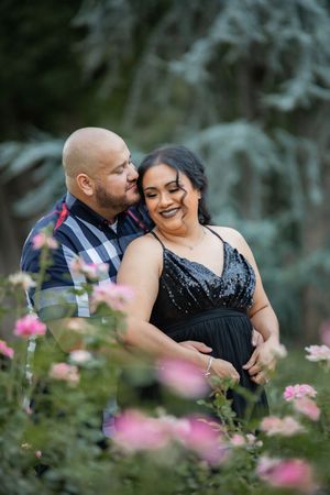 Couple embraces romantically among pink roses, woman in black sequin dress, man in plaid shirt.