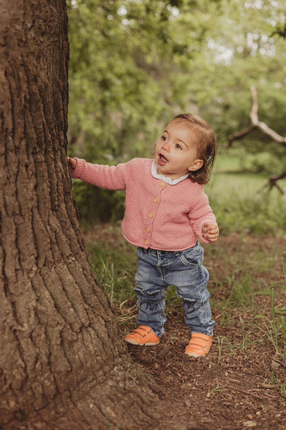 toddler in pink jumper playing hide and seek by a tree on tooting common