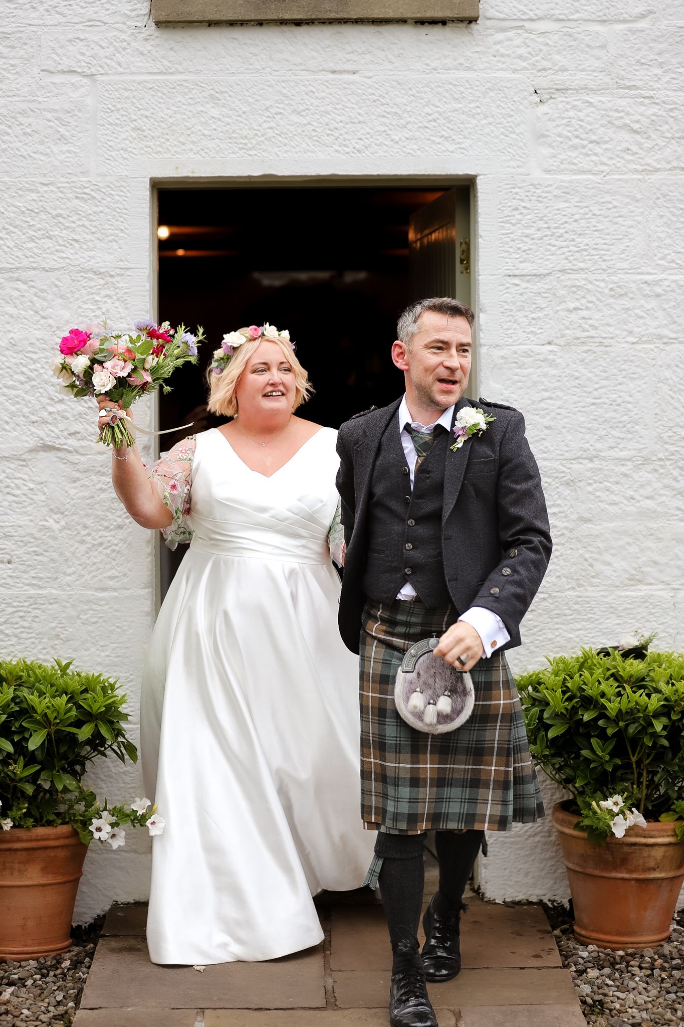 A romantic sunset wedding photo with a bride in white gown and groom wearing traditional Scottish kilt on a grassy hillside.