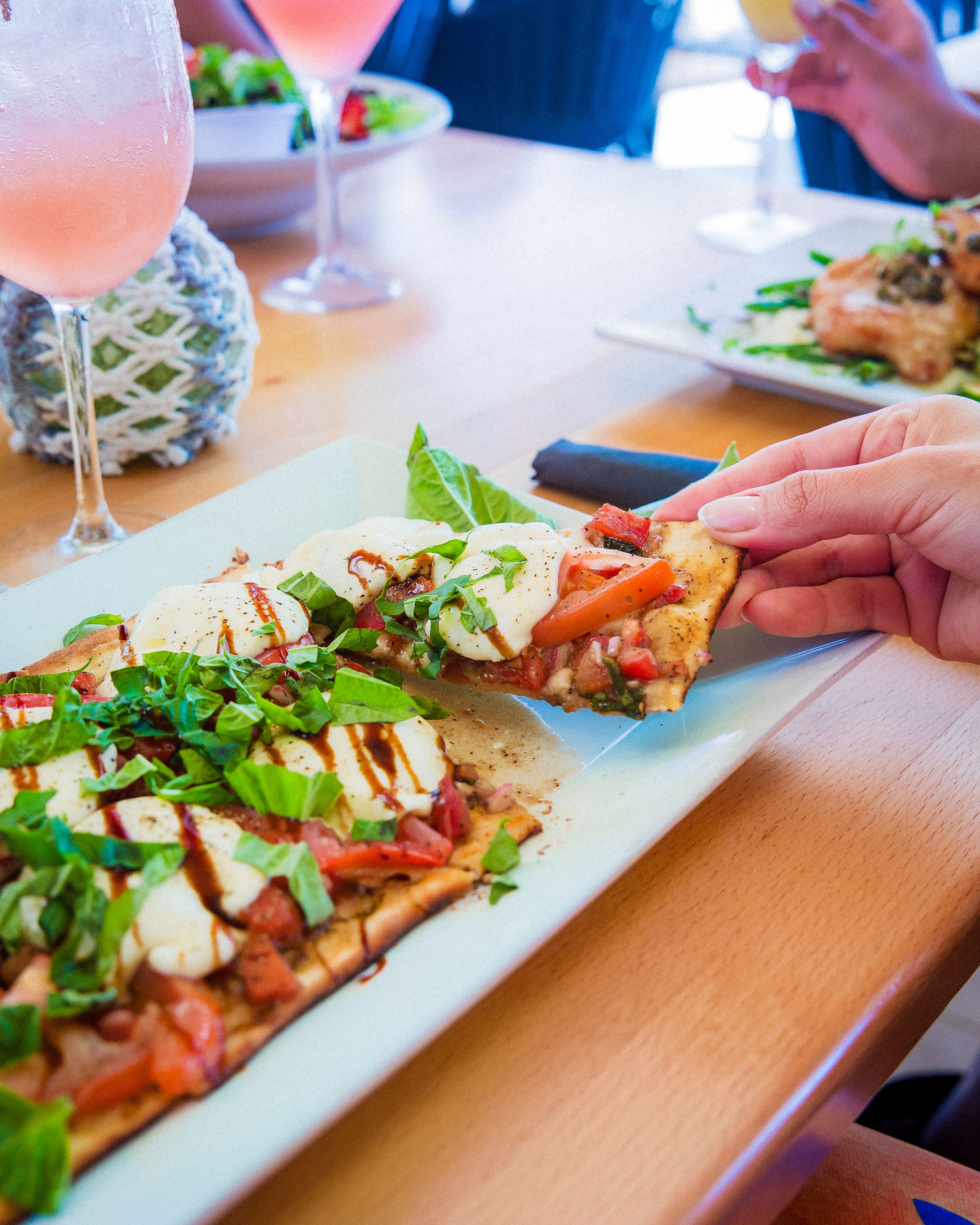 A flatbread pizza topped with fresh arugula and mozzarella sits on a white plate next to a pink cocktail at a restaurant table.