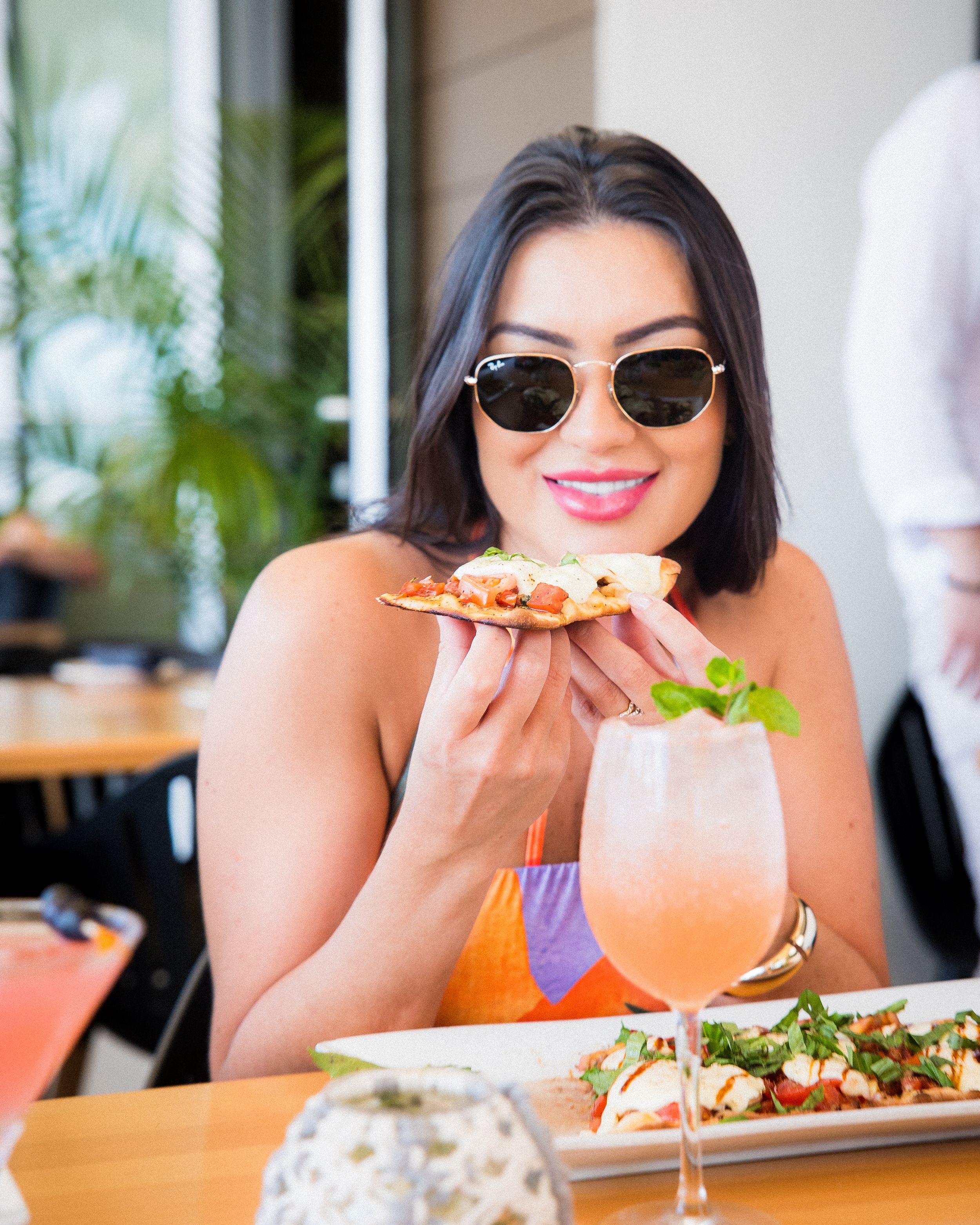 Woman in sunglasses dining at restaurant holding pizza slice with tropical cocktail and appetizers on table.