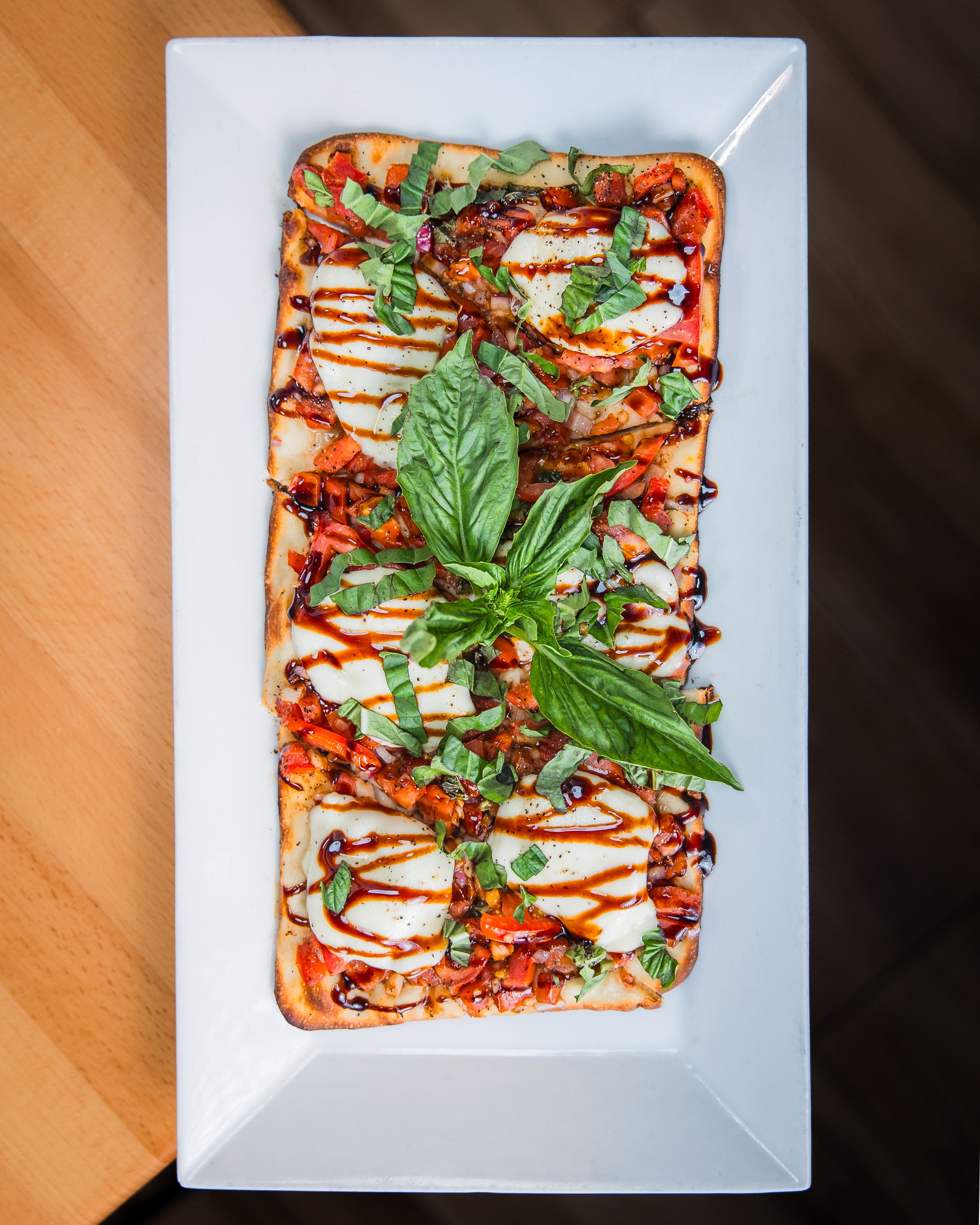 Rectangular flatbread pizza topped with mozzarella, red sauce, fresh basil leaves and balsamic drizzle served on white plate.