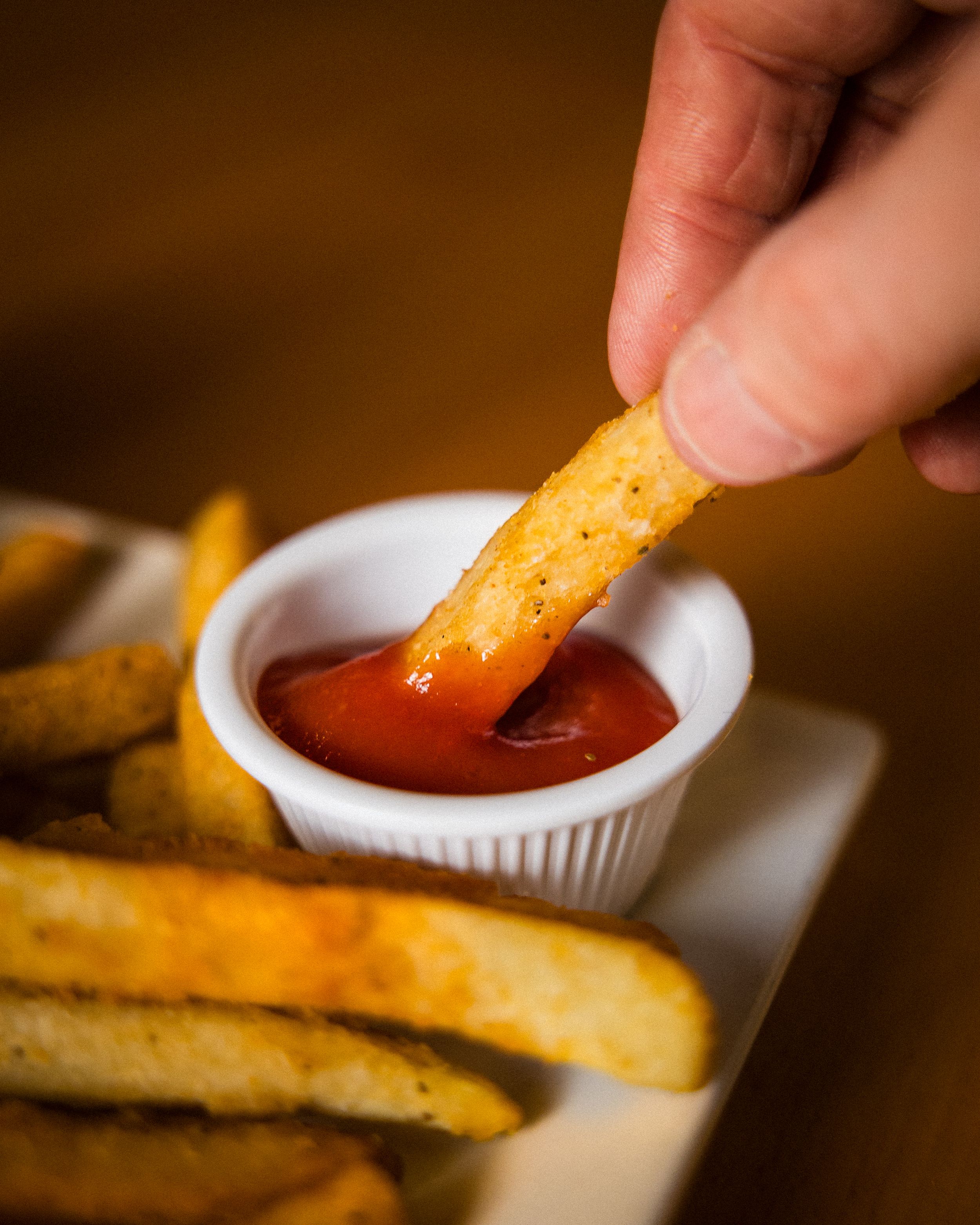 Close-up shot of hand dipping a crispy french fry into a small white ramekin of vibrant red ketchup.