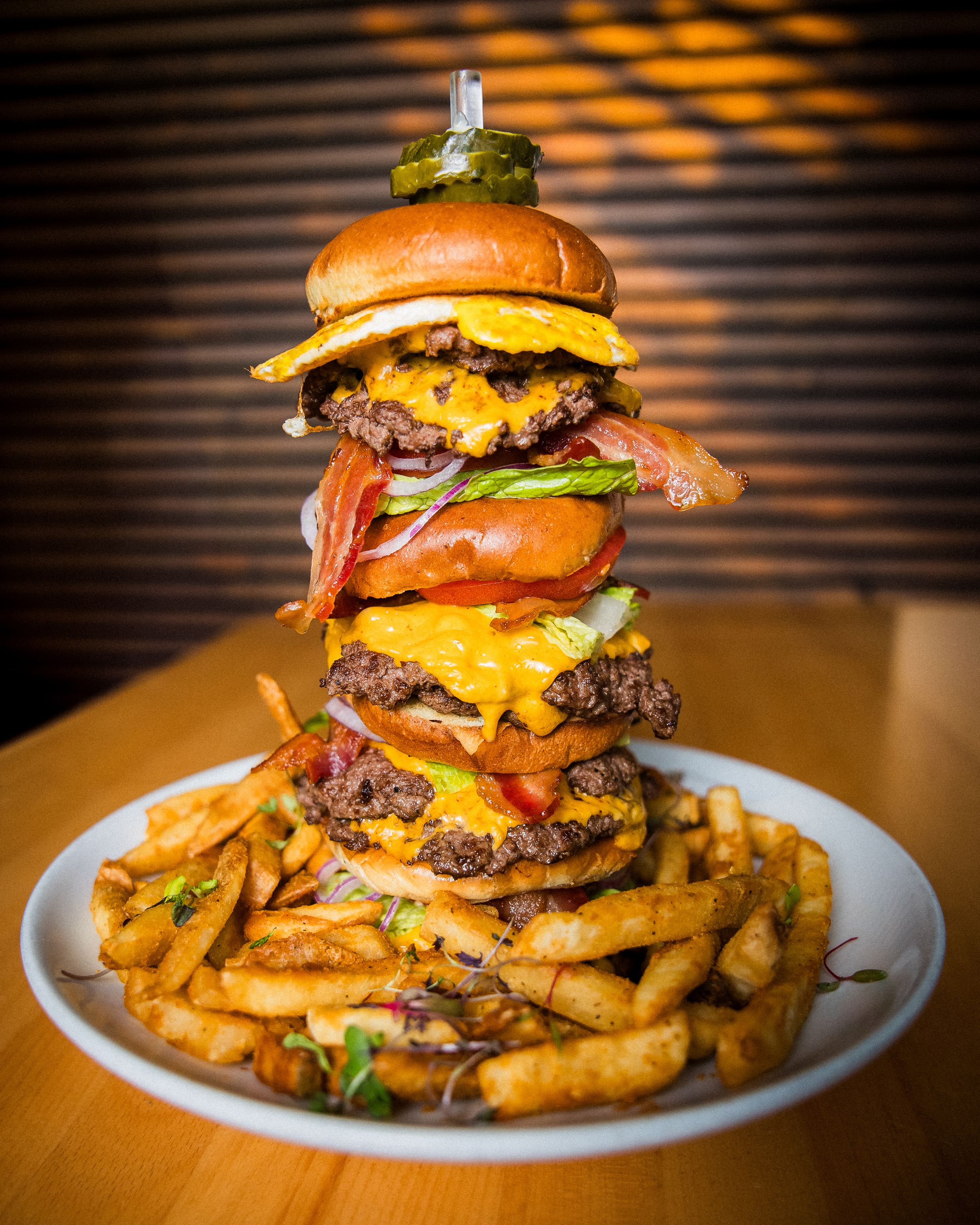 Towering triple cheeseburger with melted cheese and bacon served with crispy french fries on a white plate.