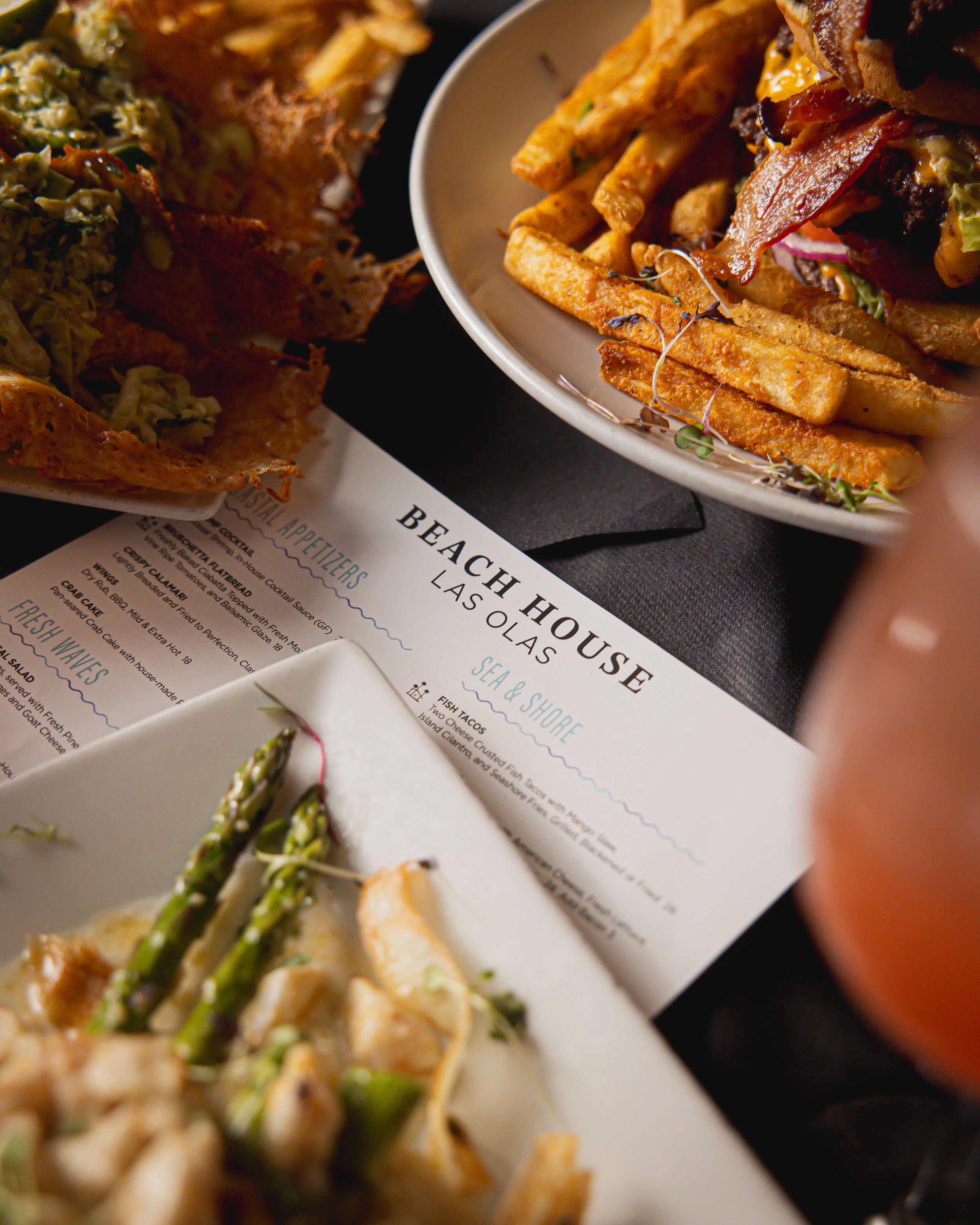 Restaurant menu card from Beach House alongside plates of french fries and asparagus dishes with cocktail in frame.