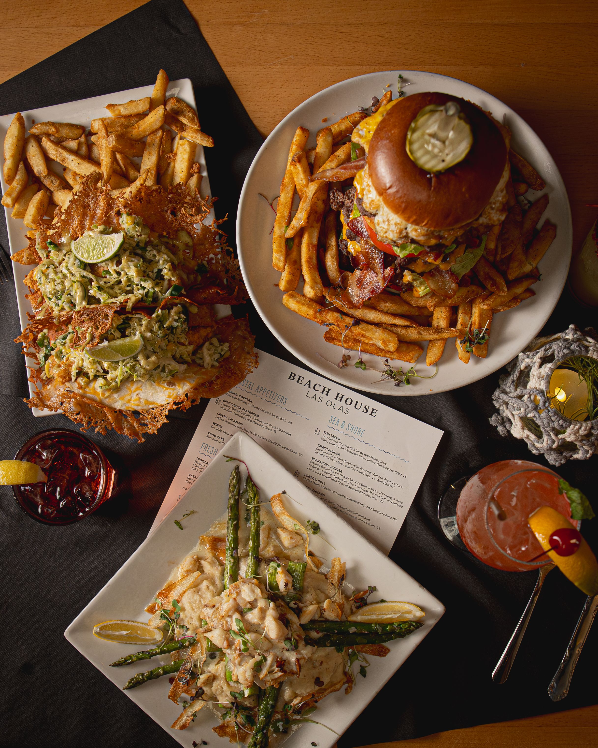 Overhead view of restaurant dishes including a loaded burger, salad with asparagus, and fries on dark surface with cocktails.