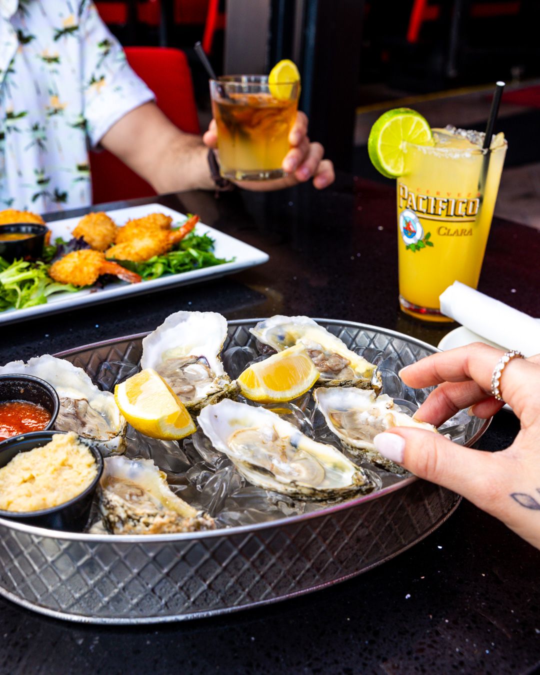 Fresh oysters on ice served with lemon wedges and sauces alongside cocktails at a restaurant table.