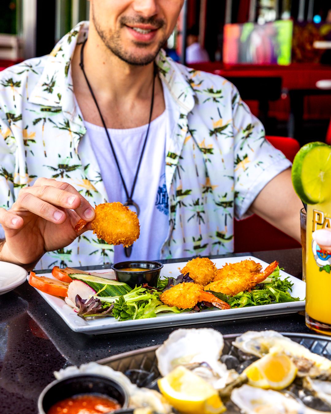 Someone in a tropical print shirt enjoys seafood and fried appetizers with raw oysters and cocktail sauce at a restaurant.
