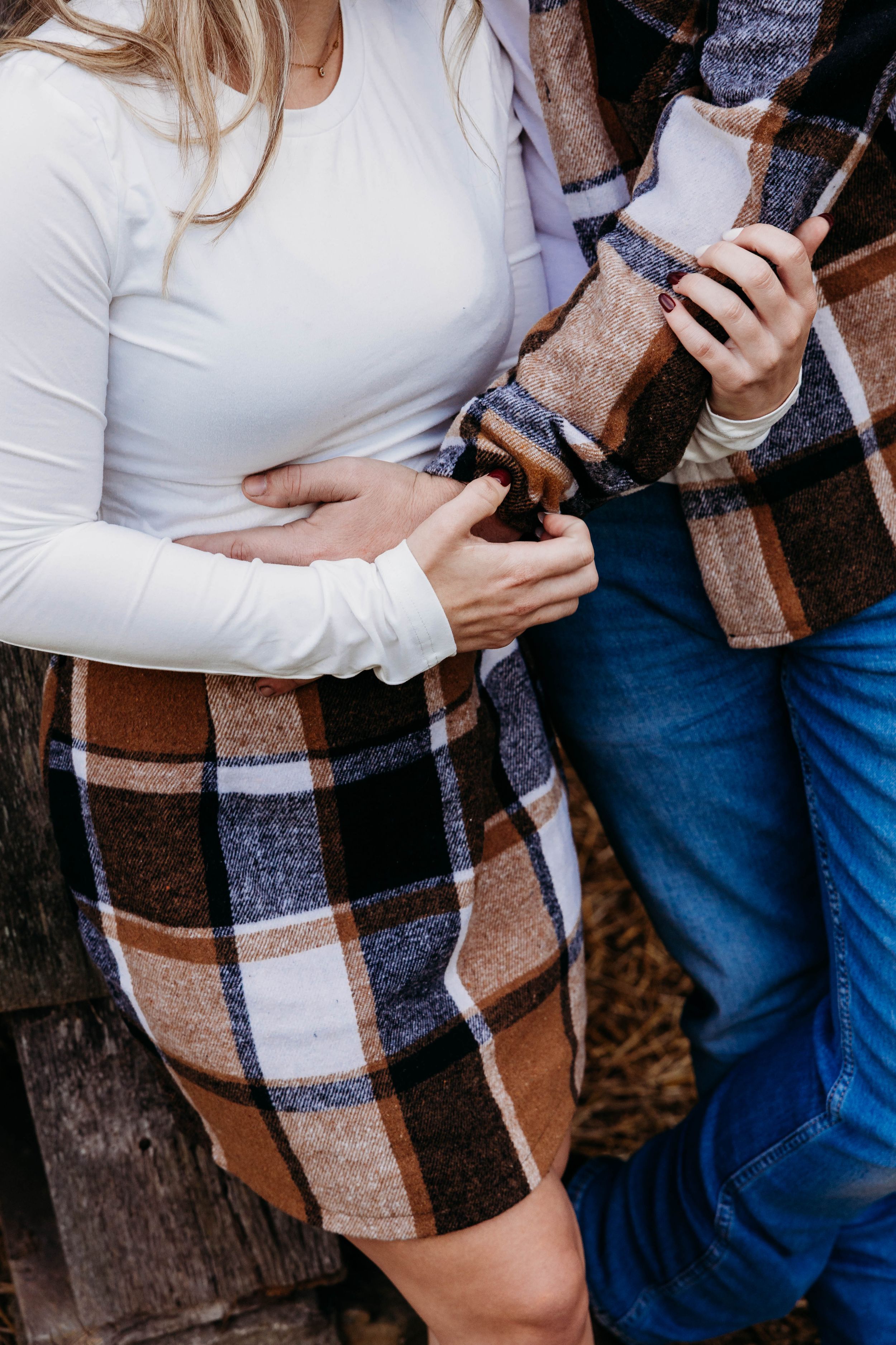 Couple wearing cozy fall outfits with plaid blanket scarf and white sweater holding phones outdoors.