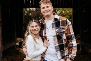 A couple in coordinated white and plaid outfits share a joyful moment together in a wooded outdoor setting.
