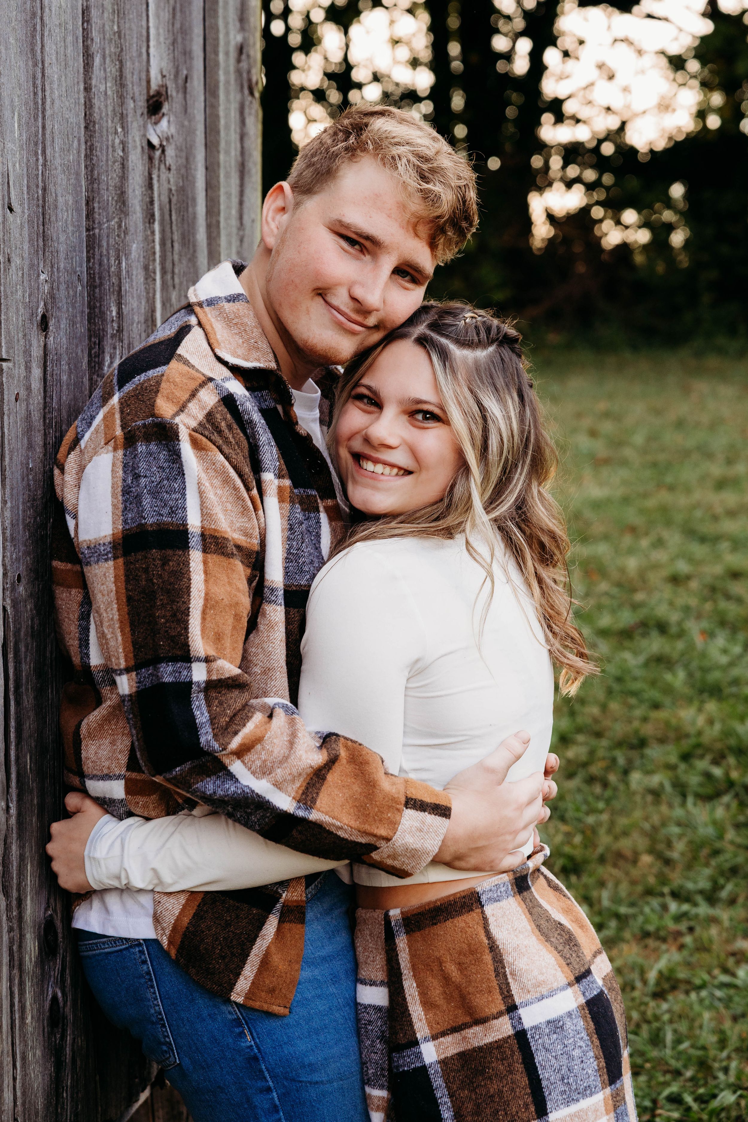 A couple embraces while leaning against a rustic wooden wall, wearing a plaid flannel shirt and white sweater at sunset.