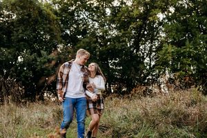 A romantic couple in casual attire walks through a sunlit meadow during a golden hour photo session.