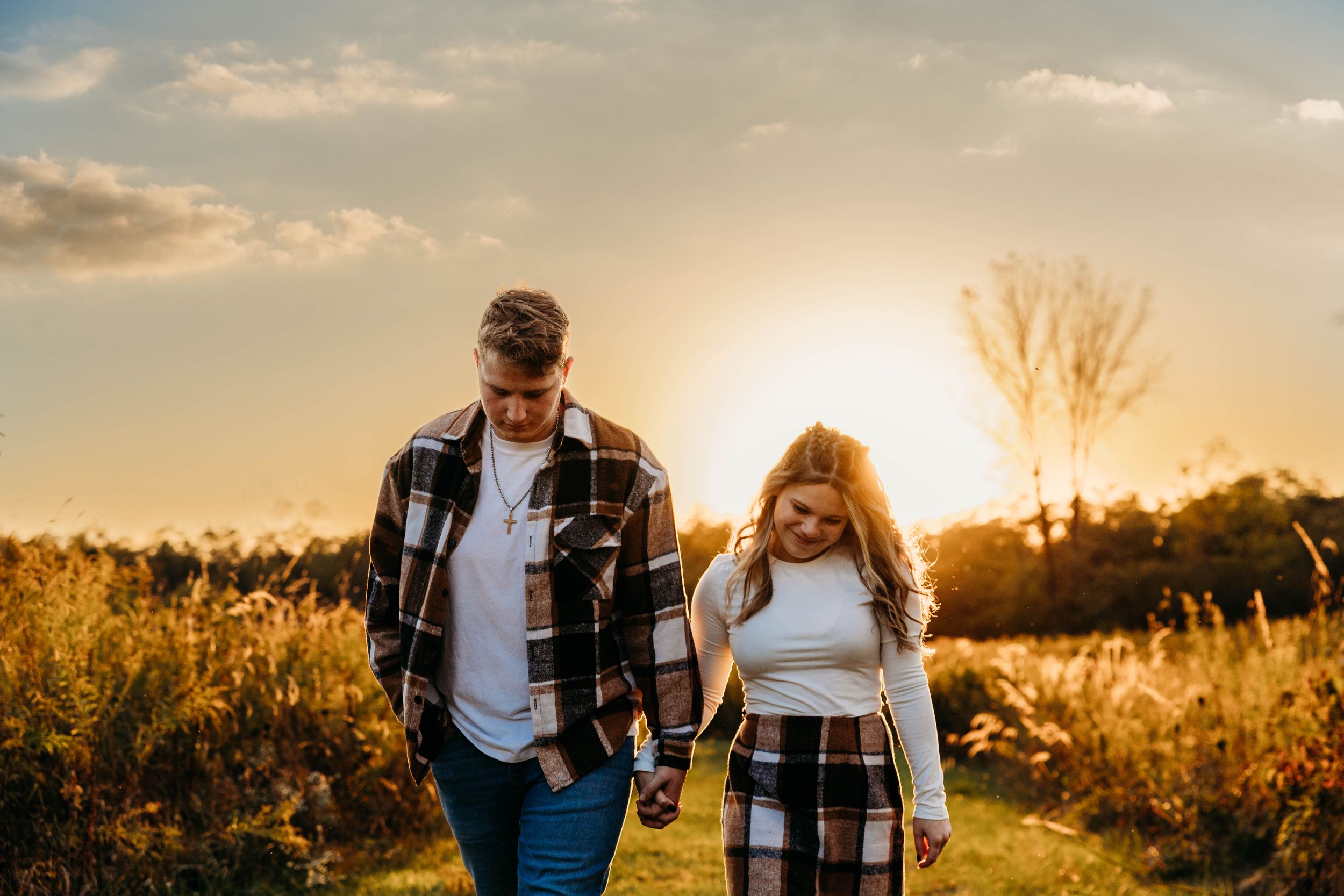 Couple walking through golden field at sunset holding hands while wearing plaid shirts and jeans.