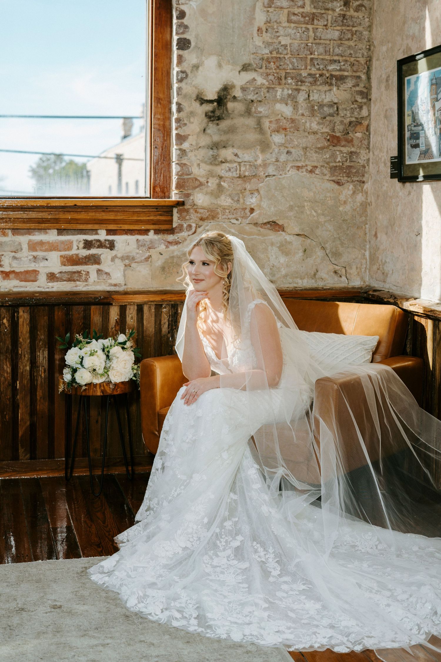 Bride in elegant lace wedding gown with flowing veil poses gracefully against rustic exposed brick wall interior.