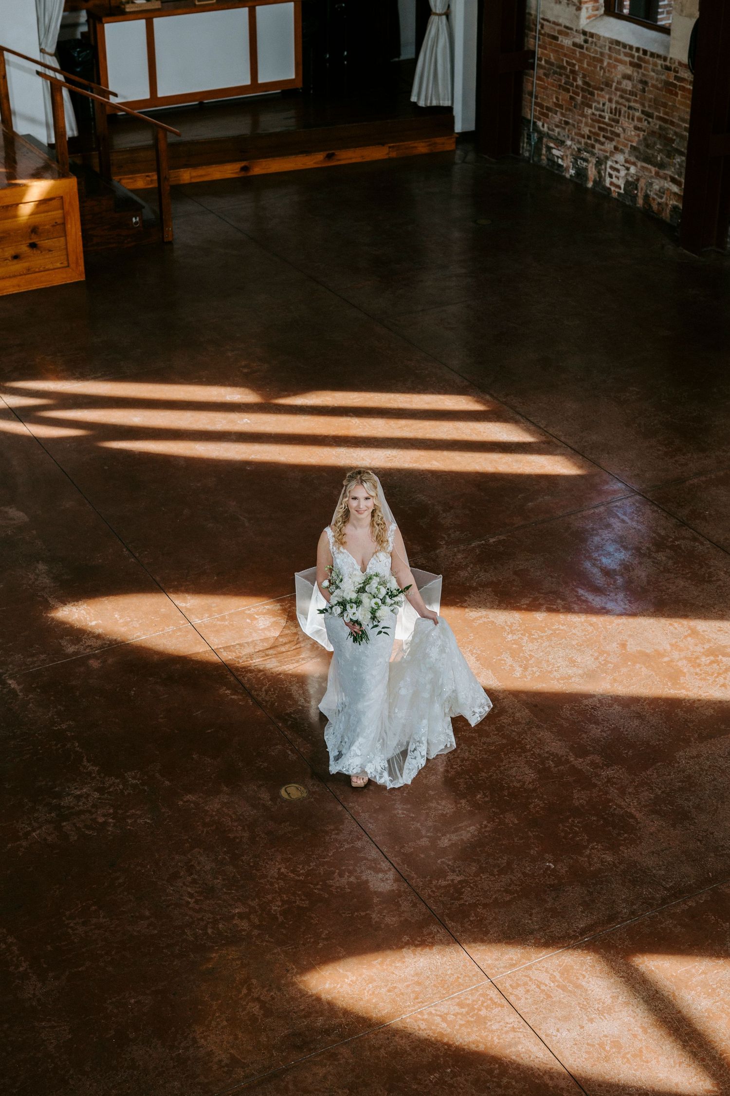 A white wedding dress lays elegantly on a dark wooden floor with dramatic natural light streaming through windows.