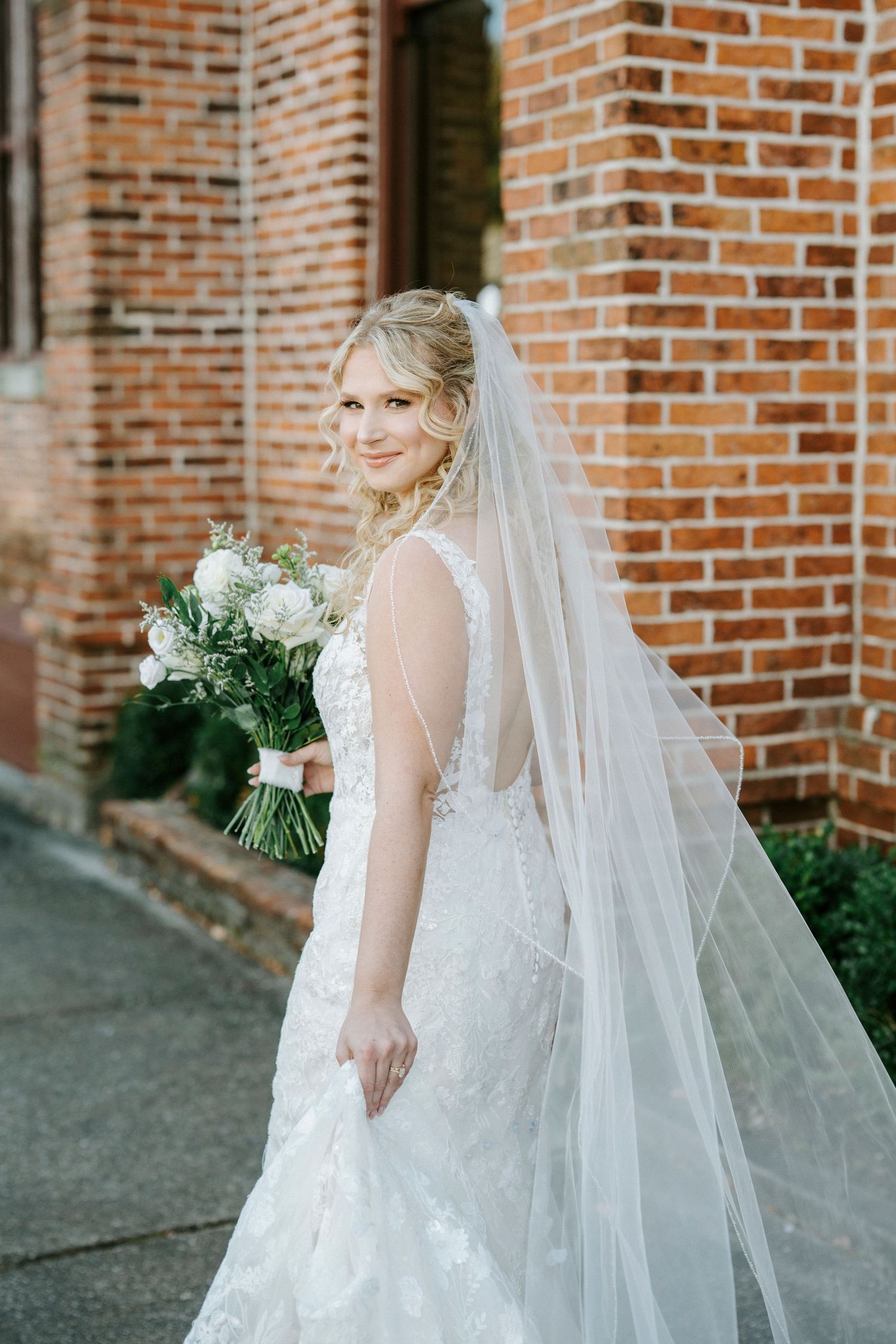 Bride in white lace wedding dress with flowing veil poses against rustic brick wall backdrop.