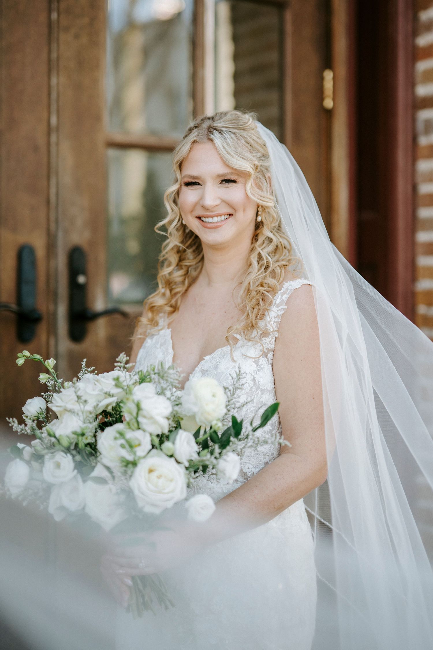 A bride in a white lace wedding dress and veil poses with an elegant white floral bouquet against a rustic brick backdrop.