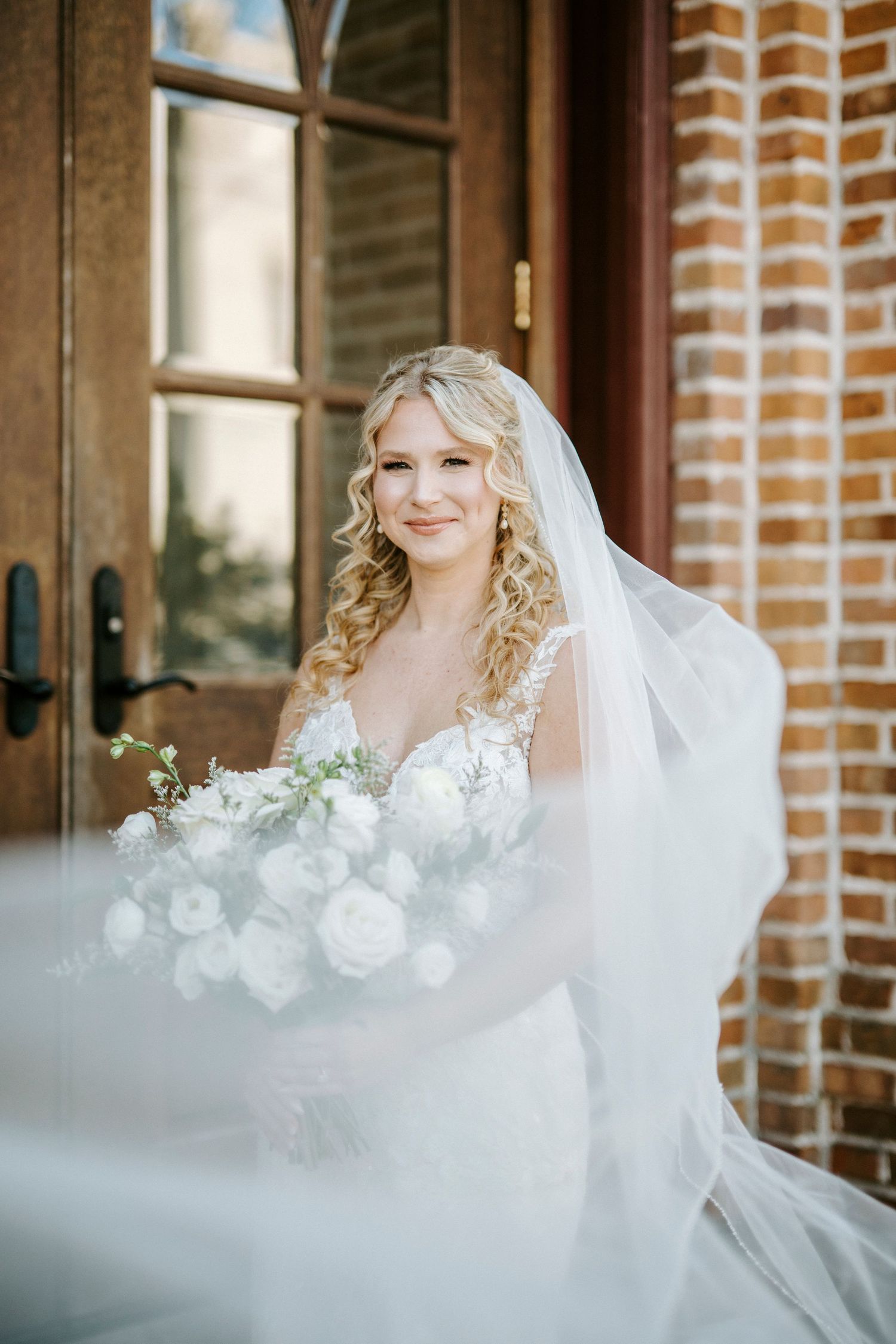 A bride in a white wedding dress with flowing veil stands by a brick building smiling on her wedding day.
