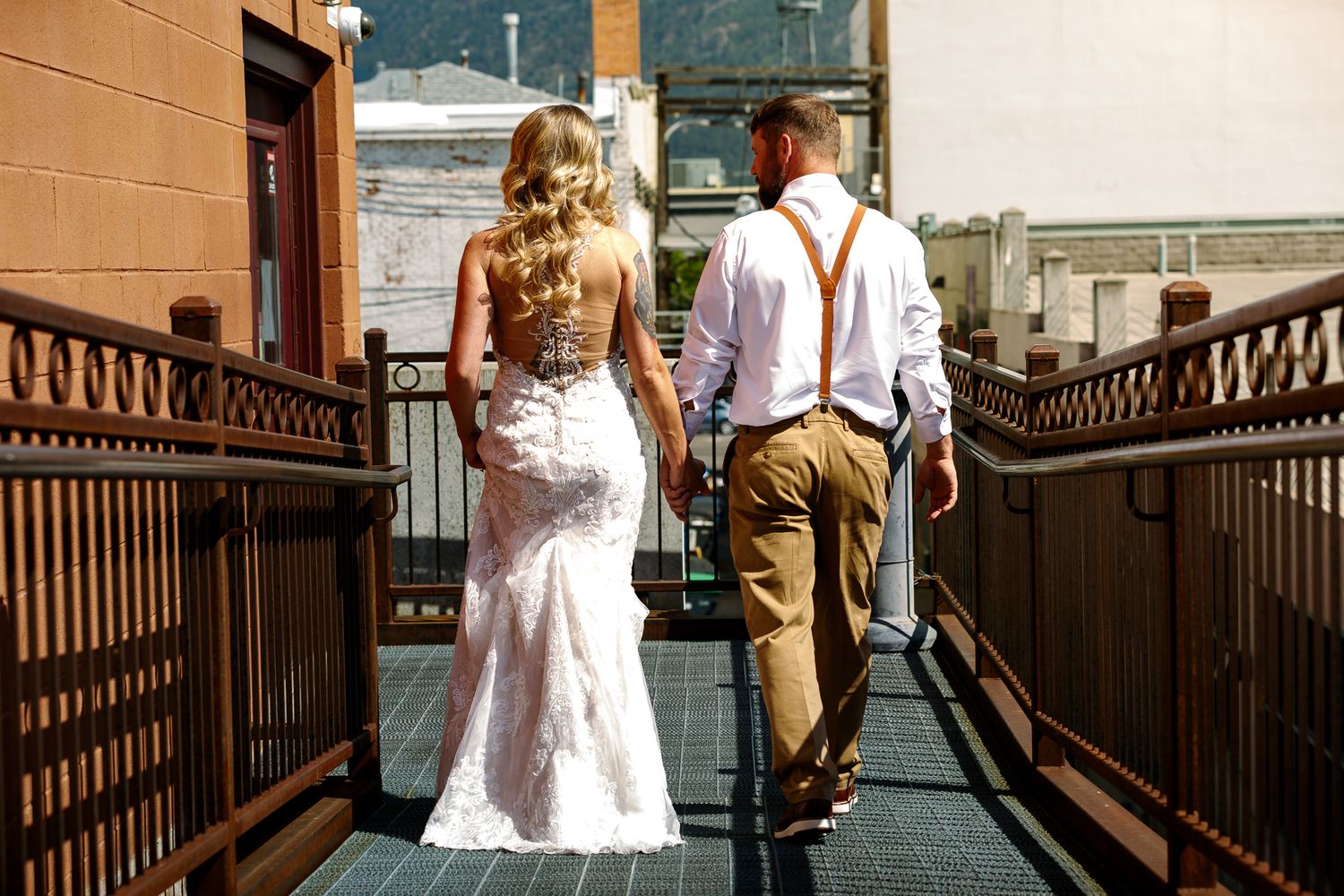 A couple walks along a rooftop deck with metal railings, the bride wearing a fitted lace gown and groom in suspenders.